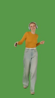 Yürürken, kulaklık takarken, kot pantolon giyerken ve turuncu bir kazak giyerken müziğin keyfini çıkaran bir insan, canlı yeşil bir arkaplan üzerine kuruludur..