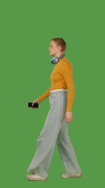 Yürümekten hoşlanan, kulaklık takan, kot pantolon giyen, turuncu bir kazak giyen, canlı yeşil ekran, krom anahtar...