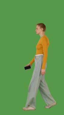 Sarı bluz ve kot pantolon giyen yeşil ekrana karşı akıllı telefonuna odaklanan biri, kroma anahtar. Yan görünüm