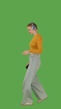 Yürümekten hoşlanan, kulaklık takan, kot pantolon giyen, turuncu kazak giyen, canlı yeşil ekran, krom anahtar, dikey görüntü,