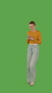 Sarı bluz ve kot pantolon giyen yeşil ekrana karşı akıllı telefonuna odaklanan biri, kroma anahtar. Yarım dönüş görünümü, dikey video