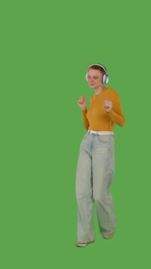 Yürümekten hoşlanan, kulaklık takan, kot pantolon giyen, turuncu kazak giyen, canlı yeşil ekran ve krom anahtarla çizilmiş bir insan. Yarım dönüş görünümü, dikey video