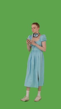 Yürürken müziği seven, kulaklık takan, kot mavisi elbise giyen, canlı yeşil ekran, krom anahtara dayanan bir insan. Yarım dönüş görünümü, dikey video