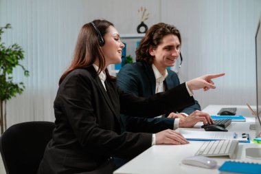 Bilgisayar başında çalışan profesyonel çağrı merkezi ajanlarından oluşan bir ekip uzman müşteri hizmetleri ve destek sunuyor. Ekranı işaret eden bir kadın meslektaşına önemli bilgiler veriyor..