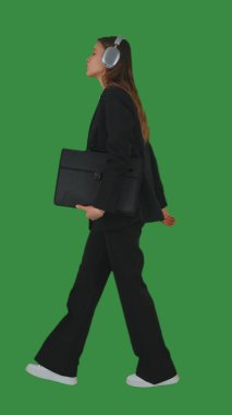 Resmi iş kıyafeti giymiş, yürüyen ve kulaklıkla müzik dinleyen bir kadın yeşil ekran ve krom anahtarla. Yan görünüm