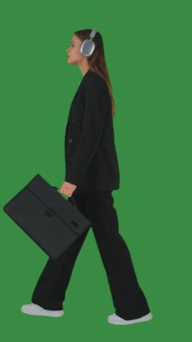 Resmi iş kıyafeti giymiş, yürüyen ve kulaklıkla müzik dinleyen bir kadın yeşil ekran ve krom anahtarla. Yan görünüm