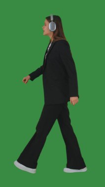 Resmi iş kıyafeti giymiş, yürüyen ve kulaklıkla müzik dinleyen bir kadın yeşil ekran ve krom anahtarla. Yan görünüm
