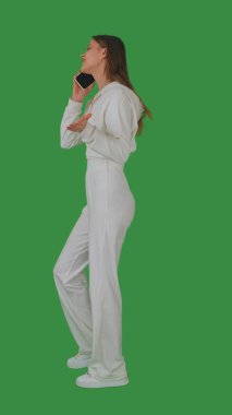 Beyaz elbiseli genç bir kadın akıllı telefonda parlak yeşil ekran, kroma anahtarıyla konuşurken yürüyor. Yan görünüm