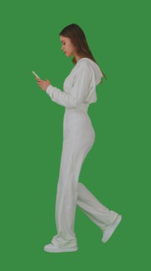 Beyaz elbiseli genç bir kadın cep telefonunu kullanırken yürüyor, yeşil ekrana karşı, krom anahtarla. Yan görünüm