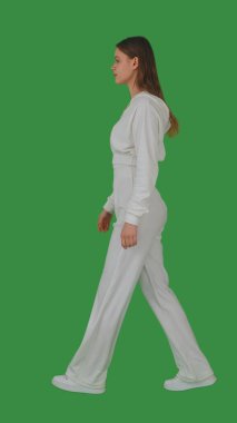 Beyaz takım elbiseli ve beyaz spor ayakkabılı bir kadın yeşil ekran ve krom anahtarla rahatça yürür. Yan görünüm