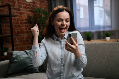 Neşeli genç bir kadın, akıllı telefonuna bakıp, iyi haberi veya başarıyı kutlarken heyecanla tezahürat yapar..