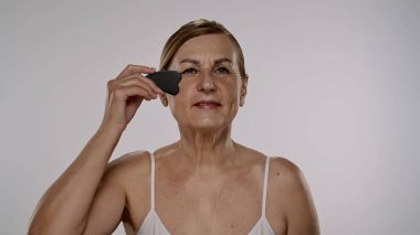 Yaşlı bir kadın yüz masajı için gua sha aracı kullanır, cilt bakımı, rahatlama ve gençleştirme teknikleri gösterir.