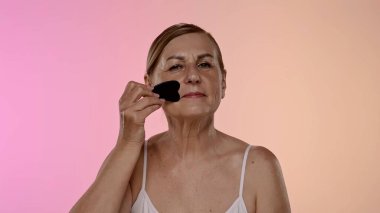 Yaşlı bir kadın yüz masajı için gua sha aracı kullanır, cilt bakımı, rahatlama ve gençleştirme teknikleri gösterir.