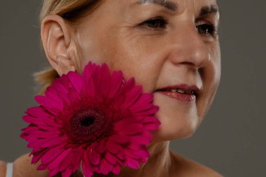 Yetişkin bir kadının yüzüne yakın sanatsal bir yaklaşım, gözlerine odaklanmış ve yanında canlı pembe bir çiçek..