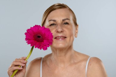 Canlı pembe bir Gerbera papatyası tutan yaşlı bir kadın. Tarafsız bir arka plana karşı güzelliği ve bilgeliği sembolize ediyor..