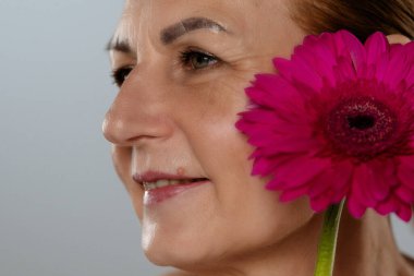 Yetişkin bir kadının yüzüne yakın sanatsal bir yaklaşım, gözlerine odaklanmış ve yanında canlı pembe bir çiçek..