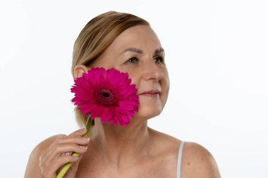 Yaşlı bir kadın canlı pembe bir Gerbera papatyası tutuyor. Beyaz bir arka plana karşı güzelliği ve bilgeliği simgeliyor..