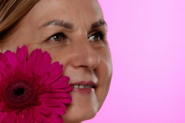Yetişkin bir kadının yüzüne yakın sanatsal bir yaklaşım, gözlerine odaklanmış ve yanında canlı pembe bir çiçek..