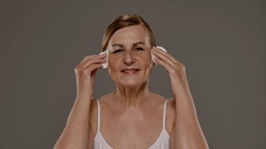 Üst düzey bir kadın yüzünü pamuk pedlerle nazikçe temizliyor, cilt bakımına ve sağlığına odaklanıyor..