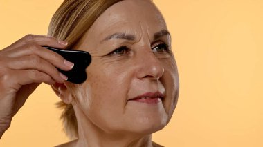 Yaşlı bir kadın yüz masajı için gua sha aleti kullanır, cilt bakımı, rahatlama ve gençleştirme teknikleri gösterir. Kapat.
