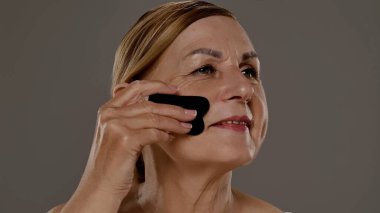 Yaşlı bir kadın yüz masajı için gua sha aracı kullanır, cilt bakımı, rahatlama ve gençleştirme teknikleri gösterir.