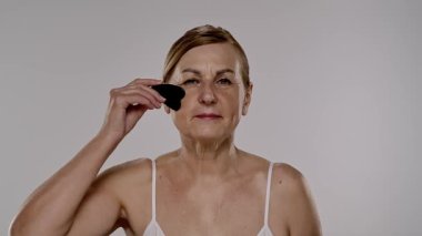Yaşlı bir kadın yüz masajı için gua sha aleti kullanır, cilt bakımı, rahatlama ve gençleştirme teknikleri gösterir. Yavaş çekim