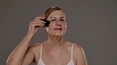 Yaşlı bir kadın yüz masajı için gua sha aleti kullanır, cilt bakımı, rahatlama ve gençleştirme teknikleri gösterir. Yavaş çekim