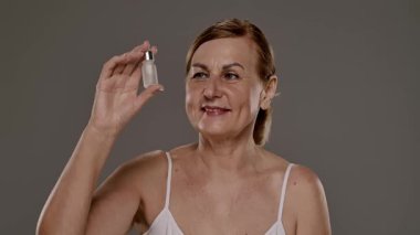 Bir kadın elinde küçük bir şişe cilt serumuyla pembe bir arka plana gülümsüyor. Yavaş çekim