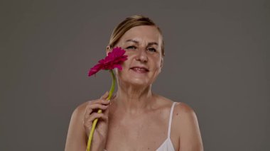 Pembe arka planda canlı pembe bir Gerbera papatyası tutan, güzelliği ve bilgeliği sembolize eden yaşlı bir kadın. Yavaş çekim