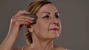 Yaşlı bir kadın yüz masajı için gua sha aleti kullanır, cilt bakımı, rahatlama ve gençleştirme teknikleri gösterir. Yaklaşın, ağır çekim.