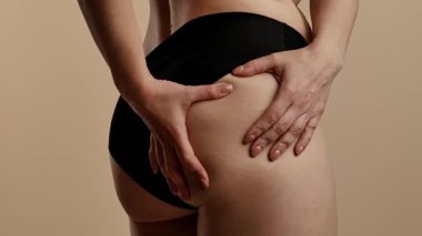 Siyah iç çamaşırlı bir kadının kalçasını çimdiklediği ve doğal vücut dokusunu ön plana çıkardığı yakın plan bir görüntü. Görüntü, yumuşak ve nötr bir arkaplana karşı çekiliyor. Yavaş çekim