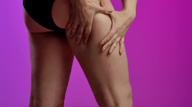 Siyah iç çamaşırlı bir kadının kalçasını çimdiklediği ve doğal vücut dokusunu ön plana çıkardığı yakın plan bir görüntü. Görüntü yumuşak mor bir arkaplan üzerinde yakalanır. Yavaş çekim