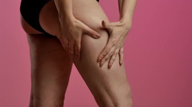 Siyah iç çamaşırlı bir kadının kalçasını çimdiklediği ve doğal vücut dokusunu ön plana çıkardığı yakın plan bir görüntü. Görüntü, yumuşak pembe bir arkaplana karşı çekiliyor. Yavaş çekim