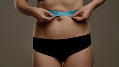 Bir kadın belini bir mezurayla ölçer, spor süreçlerini ve genel sağlık hedeflerini takip etmeye odaklanır. Yaklaşın, ağır çekim.