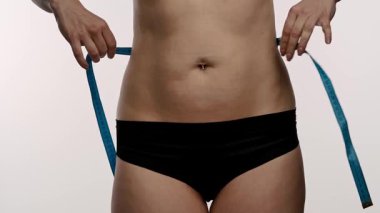 Siyah iç çamaşırlı bir kadın bel ölçüsünü mavi bir bantla ölçer, spor ve sağlık hedeflerini sembolize eder. Yaklaşın, ağır çekim.