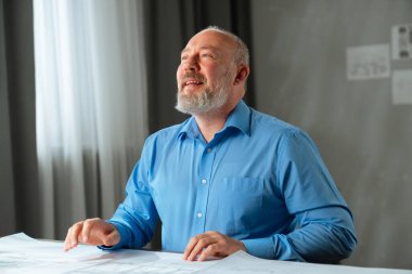 Mavi gömlekli bir mimar arka plana bakar ve gülümser. Evlerin ve dairelerin modellerinin olduğu çizimler masanın üzerine yayılmış durumda. Arka planda duvar planları ve yumuşak ışıklandırma var..