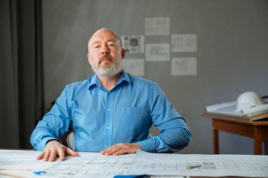 Mavi gömlekli bir mimar kendinden emin bir şekilde kameraya bakar. Evlerin ve dairelerin yerleşim planlarının olduğu çizimler masanın üzerine yayılmış..