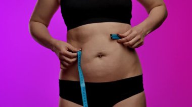 Siyah iç çamaşırlı bir kadın bel ölçüsünü mavi bir bantla ölçer, spor ve sağlık hedeflerini sembolize eder. Yaklaşın, ağır çekim.