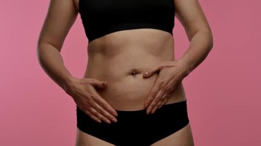 Görüntüde, göbeğinde kalp şekli oluşturan bir kadın, pembe bir arka plana karşı aşkı ya da sağlık bilincini sembolize ediyor. Yaklaşın, ağır çekim.