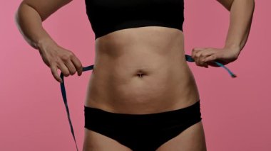 Siyah iç çamaşırlı bir kadın bel ölçüsünü mavi bir bantla ölçer, spor ve sağlık hedeflerini sembolize eder. Yaklaşın, ağır çekim.