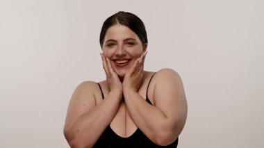 Siyah saçlı neşeli bir kadın gülümserken yüzüne dokunuyor. Arka plan beyaz renkte. Yavaş çekim