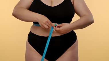 Bir kadın belini bir mezurayla ölçer, spor süreçlerini ve genel sağlık hedeflerini takip etmeye odaklanır. Yavaş çekim