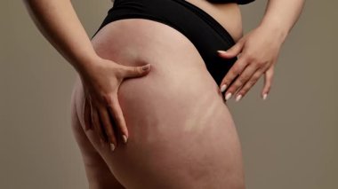 Siyah iç çamaşırlı bir kadının kalçasını çimdiklediği ve doğal vücut dokusunu ön plana çıkardığı yakın plan bir görüntü. Görüntü, yumuşak ve nötr bir arkaplana karşı çekiliyor. Yavaş çekim