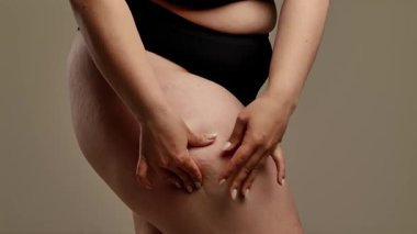 Siyah iç çamaşırlı bir kadının kalçasını çimdiklediği ve doğal vücut dokusunu ön plana çıkardığı yakın plan bir görüntü. Görüntü, yumuşak ve nötr bir arkaplana karşı çekiliyor. Yavaş çekim