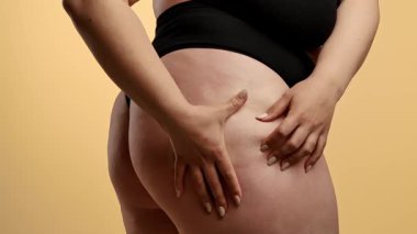 Siyah iç çamaşırlı bir kadının kalçasını çimdiklediği ve doğal vücut dokusunu ön plana çıkardığı yakın plan bir görüntü. Görüntü, yumuşak ve nötr bir arkaplana karşı çekiliyor. Yavaş çekim