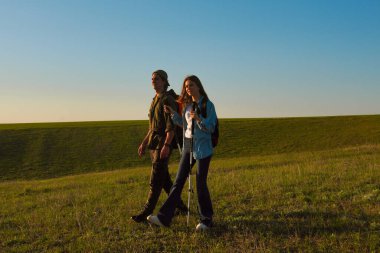 Genç bir çift, günbatımında yürüyüş sırıkları ve sırt çantalarıyla uçsuz bucaksız yeşil bir alanda doğa yürüyüşü yapıyor..