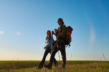 Genç bir çift, günbatımında yürüyüş sırıkları ve sırt çantalarıyla uçsuz bucaksız yeşil bir alanda doğa yürüyüşü yapıyor..