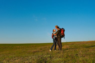 Bir çift, açık yeşil çayırlarda, açık mavi gökyüzünün altında, aşkı ve macerayı simgeleyen bir yürüyüş sırasında kucaklaşır..
