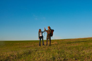 İki yürüyüşçü açık mavi gökyüzünün altında yeşil bir alanda beşlik çakar, bir başarı ve neşe anlarını paylaşırlar..