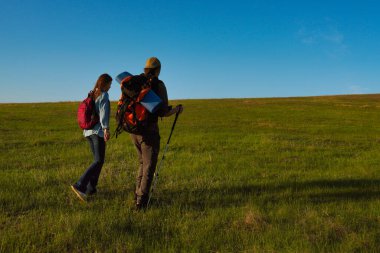Genç bir çift, günbatımında yürüyüş sırıkları ve sırt çantalarıyla uçsuz bucaksız yeşil bir alanda doğa yürüyüşü yapıyor..